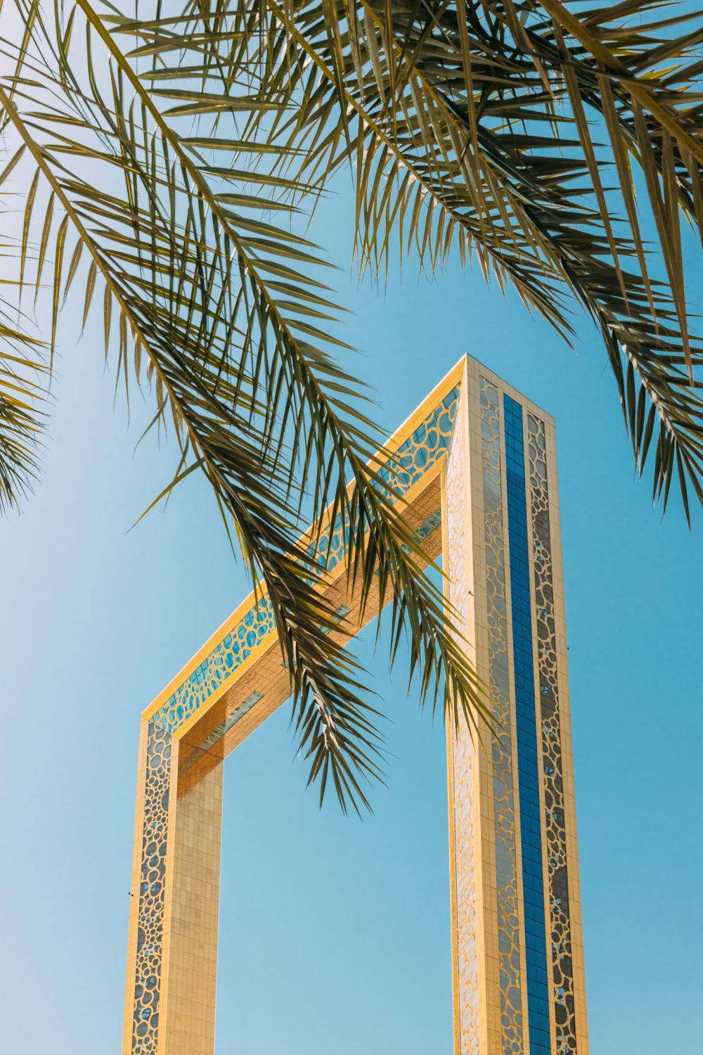 Vertical High quality photo. View Of Dubai Frame On Sunny Blue Sky Under Palm Branches. Dubai Frame Is Architectural Landmark In Zabeel Park, Dubai. It Holds Record For Largest Frame In The World. Travel Landmark Of UAE.