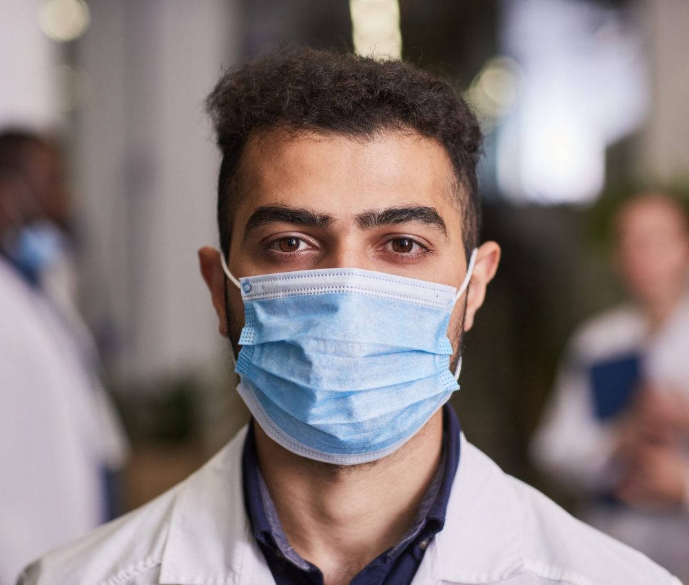 Young contemporary healthcare worker in mask and lab coat standing against his colleague having meeting in hospital corridor